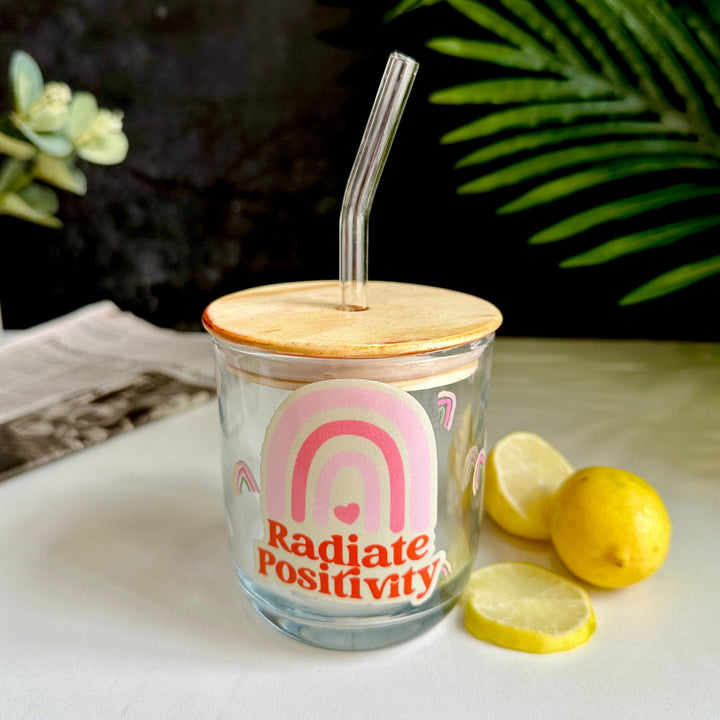 Glass tumbler with wooden lid and straw, featuring a 'Radiate Positivity' design, on a surface with lemons and greenery.